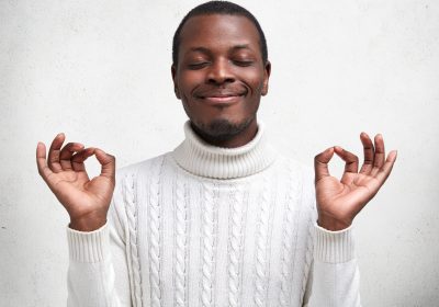 Content dark skinned young male keeps eyes shut and fingers in mudra sign, has calm expression, tries to relax after tired day, wears white sweater, poses against concrete studio background.